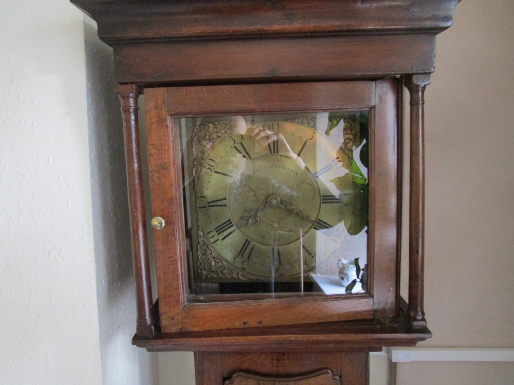An antique dark oak longcase clock featuring a glazed hood with brass dial, slim trunk door, and plinth base. View II