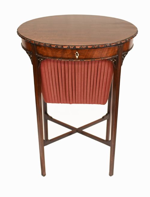 fig 1. An Edwardian mahogany sewing table featuring a carved top rim, a central keyhole escutcheon, a pleated red fabric basket underneath, and tapered legs connected by an X-frame stretcher.