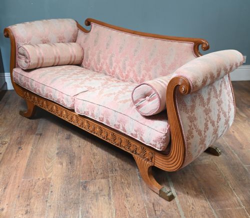 fig 2. A Regency carved walnut sofa featuring scrolled arms, anthemion leaf relief carving along the apron frieze, pink patterned upholstery, and matching bolster cushions.