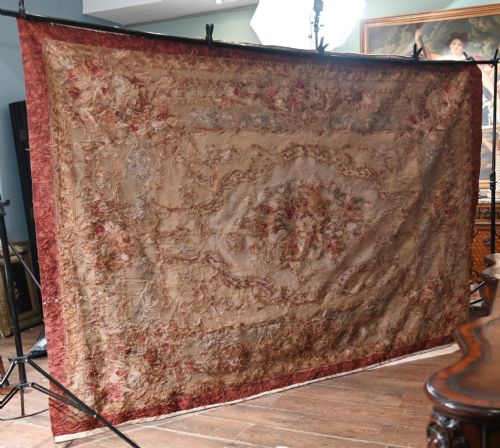 fig 1. A large rectangular French Aubusson style flat-weave tapestry featuring a central floral bouquet medallion surrounded by scrolling foliate borders in pink, beige, and muted green shades.