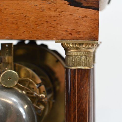 fig 1. A French Charles X portico clock crafted from dark brown inlaid wood with four fluted columns, gilt bronze accents, a white enamel dial with Roman numerals, and a hanging brass pendulum.