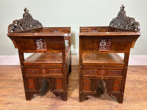 fig 1. A pair of warm brown wooden armchairs featuring elaborate carved openwork crestings and geometric mother-of-pearl inlays across the seats and panels.