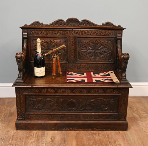 fig 1. A Victorian dark oak carved settle featuring carved lion head armrests, detailed floral and geometric back panels, and a lift-up storage seat.