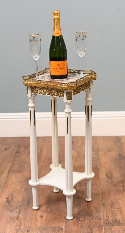 fig 1. A white painted tall French pedestal stand featuring four fluted legs, gilt brass mounts, a lower tier, and a shaped variegated marble top with a brass gallery.