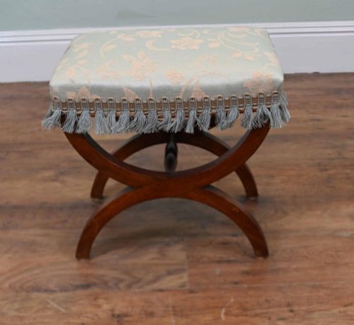 fig 1. A Regency style mahogany stool featuring curved x-frame legs, a central turned stretcher, and a pale blue patterned damask upholstered seat finished with grey fringe trim.