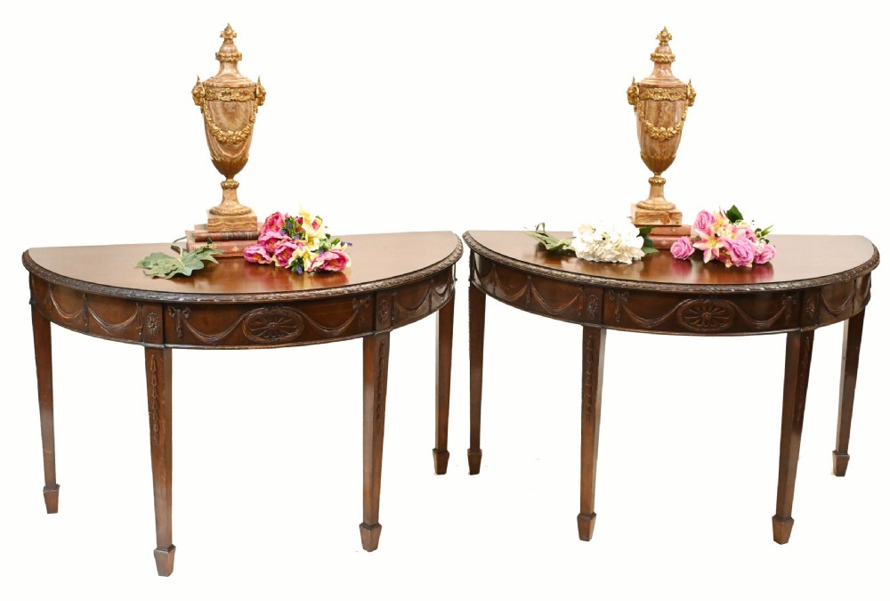 A pair of demilune mahogany console tables featuring carved friezes with decorative swags and oval paterae, supported by tapered square legs. View II