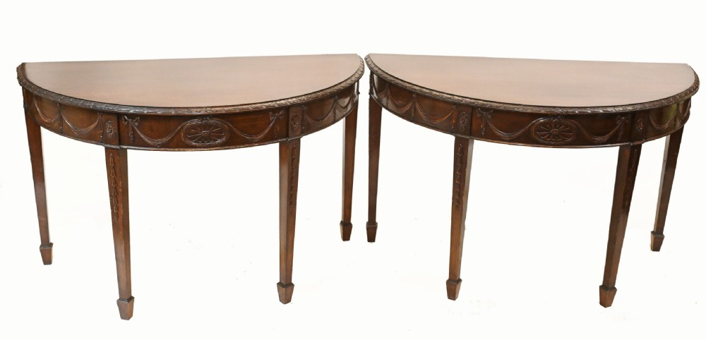 A pair of demilune mahogany console tables featuring carved friezes with decorative swags and oval paterae, supported by tapered square legs.