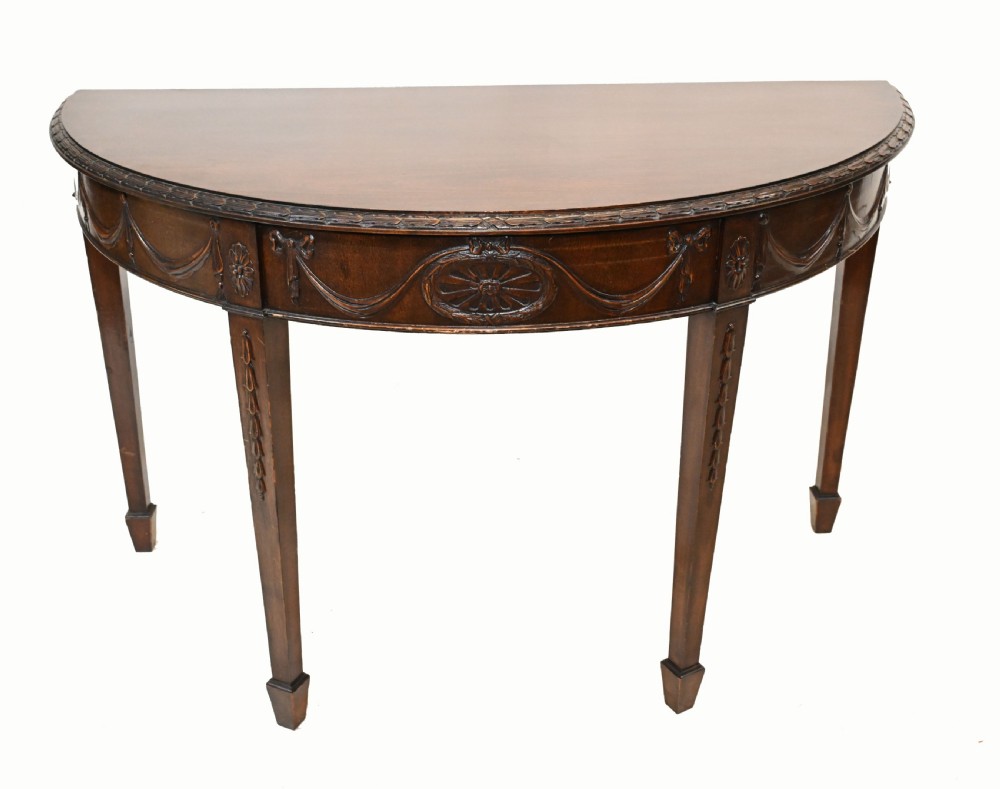 A pair of demilune mahogany console tables featuring carved friezes with decorative swags and oval paterae, supported by tapered square legs. View IV
