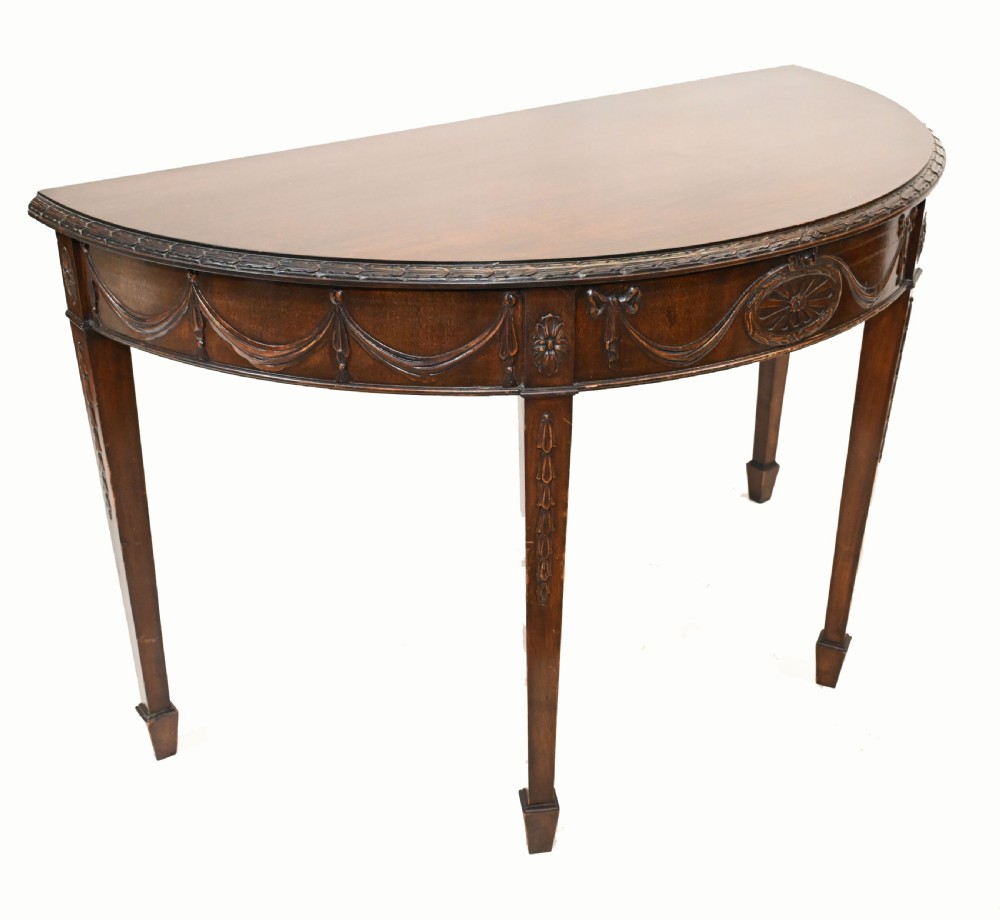 A pair of demilune mahogany console tables featuring carved friezes with decorative swags and oval paterae, supported by tapered square legs. View V