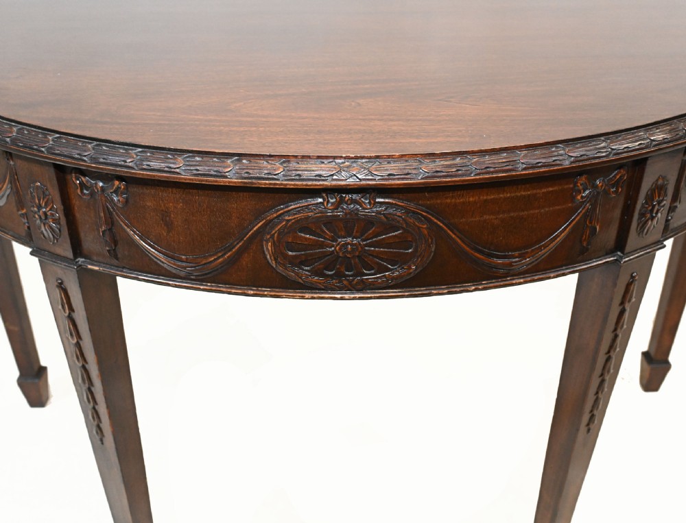 A pair of demilune mahogany console tables featuring carved friezes with decorative swags and oval paterae, supported by tapered square legs. View VI