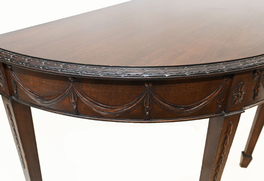 A pair of demilune mahogany console tables featuring carved friezes with decorative swags and oval paterae, supported by tapered square legs. View VII