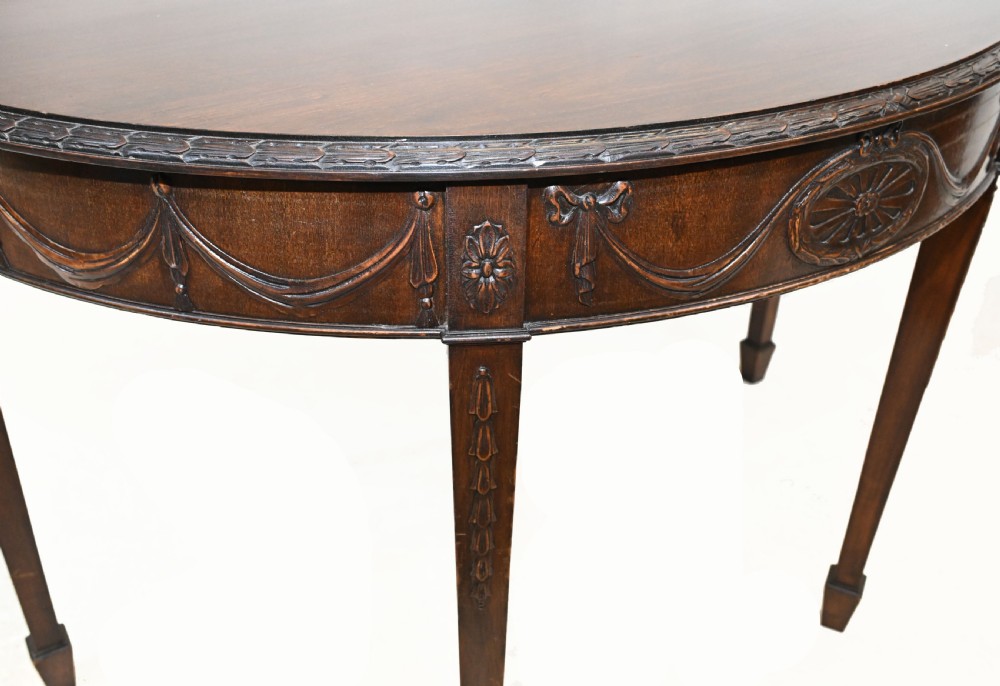 A pair of demilune mahogany console tables featuring carved friezes with decorative swags and oval paterae, supported by tapered square legs. View VIII