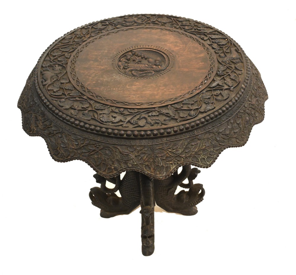 A circular dark carved teak side table featuring dense floral relief carving on the top and apron supported by a tripod base.