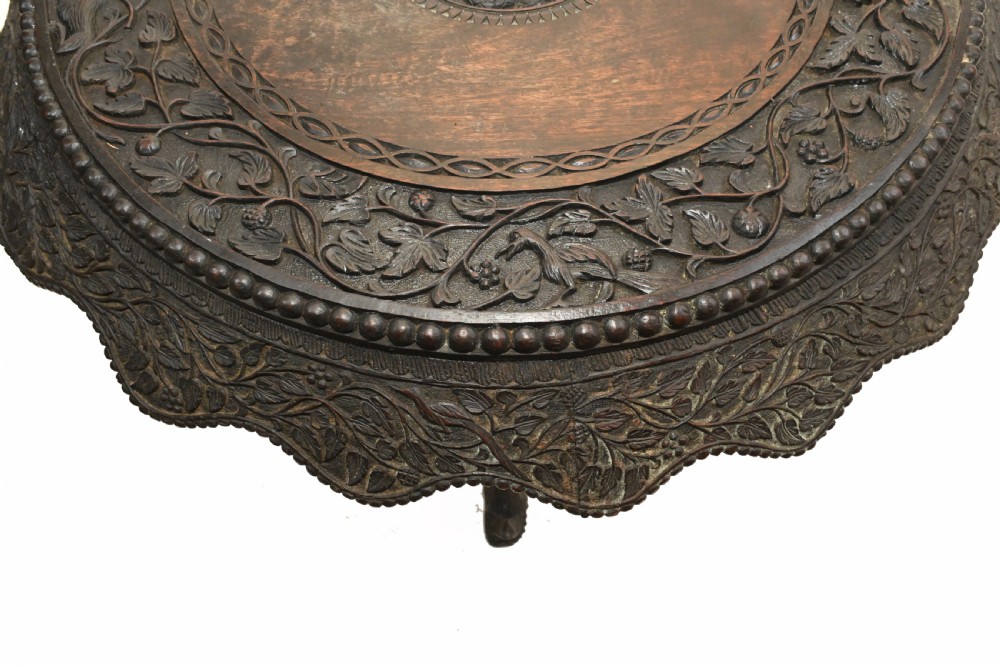 A circular dark carved teak side table featuring dense floral relief carving on the top and apron supported by a tripod base. View III