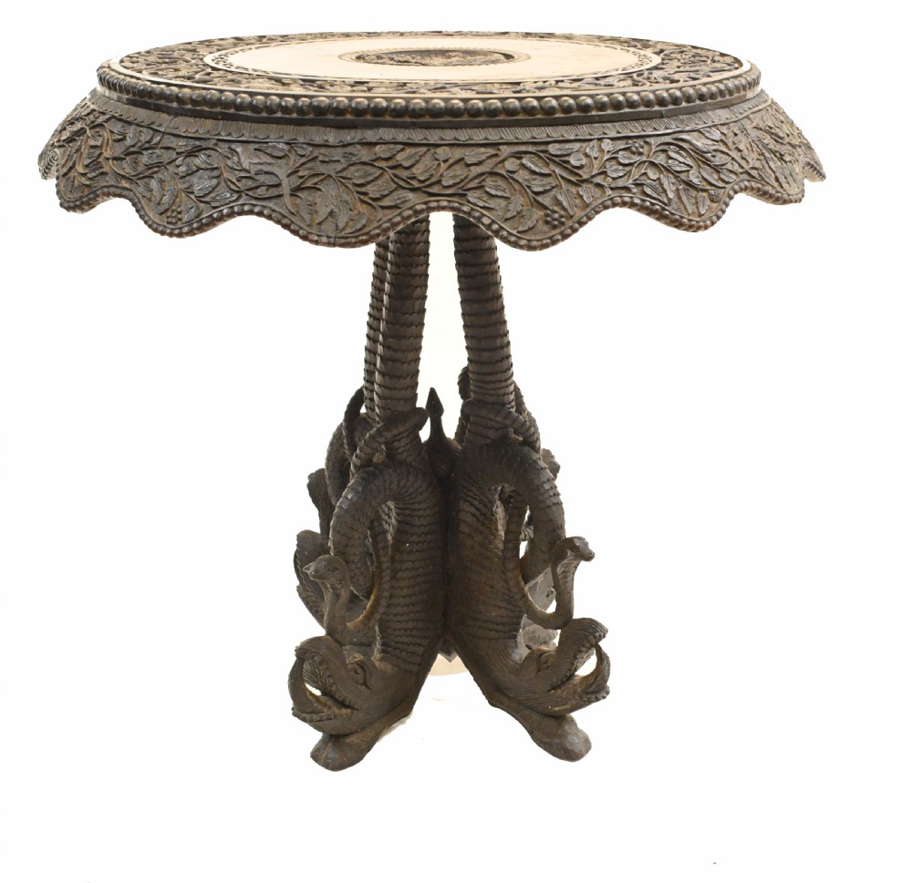 A circular dark carved teak side table featuring dense floral relief carving on the top and apron supported by a tripod base. View VII