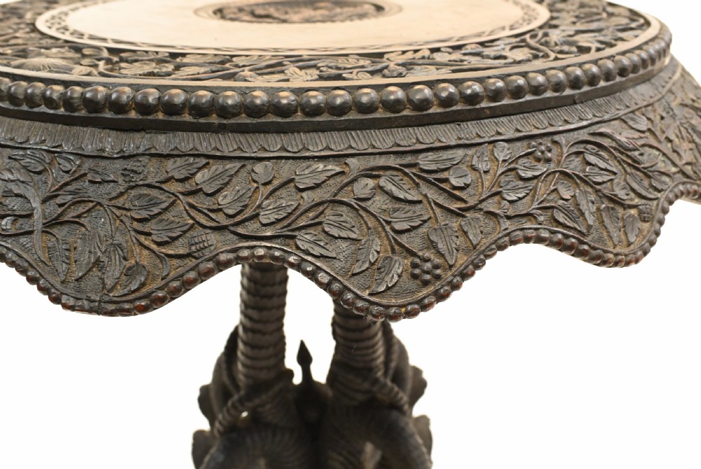 A circular dark carved teak side table featuring dense floral relief carving on the top and apron supported by a tripod base. View VIII