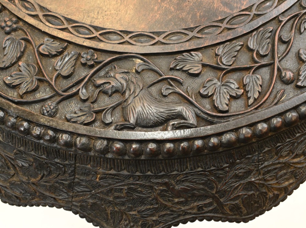 A circular dark carved teak side table featuring dense floral relief carving on the top and apron supported by a tripod base. View X