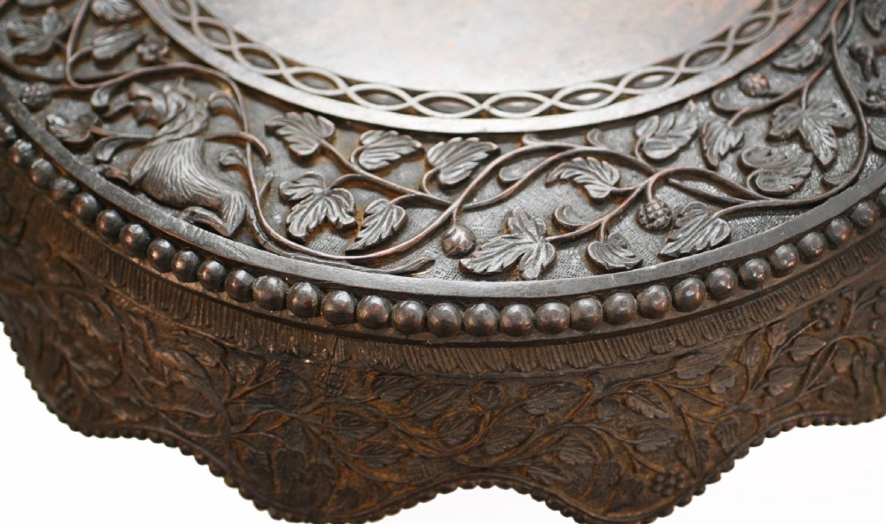 A circular dark carved teak side table featuring dense floral relief carving on the top and apron supported by a tripod base. View XII