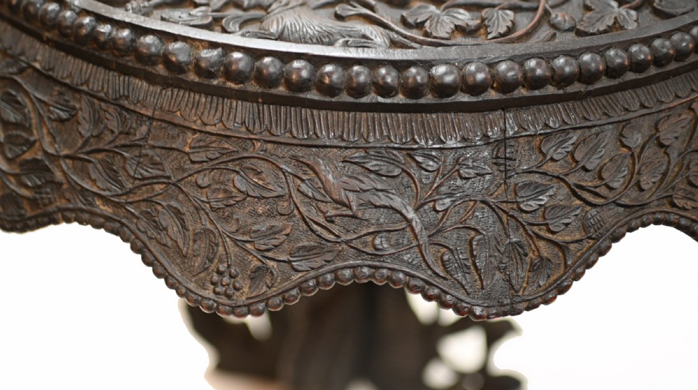 A circular dark carved teak side table featuring dense floral relief carving on the top and apron supported by a tripod base. View XIII