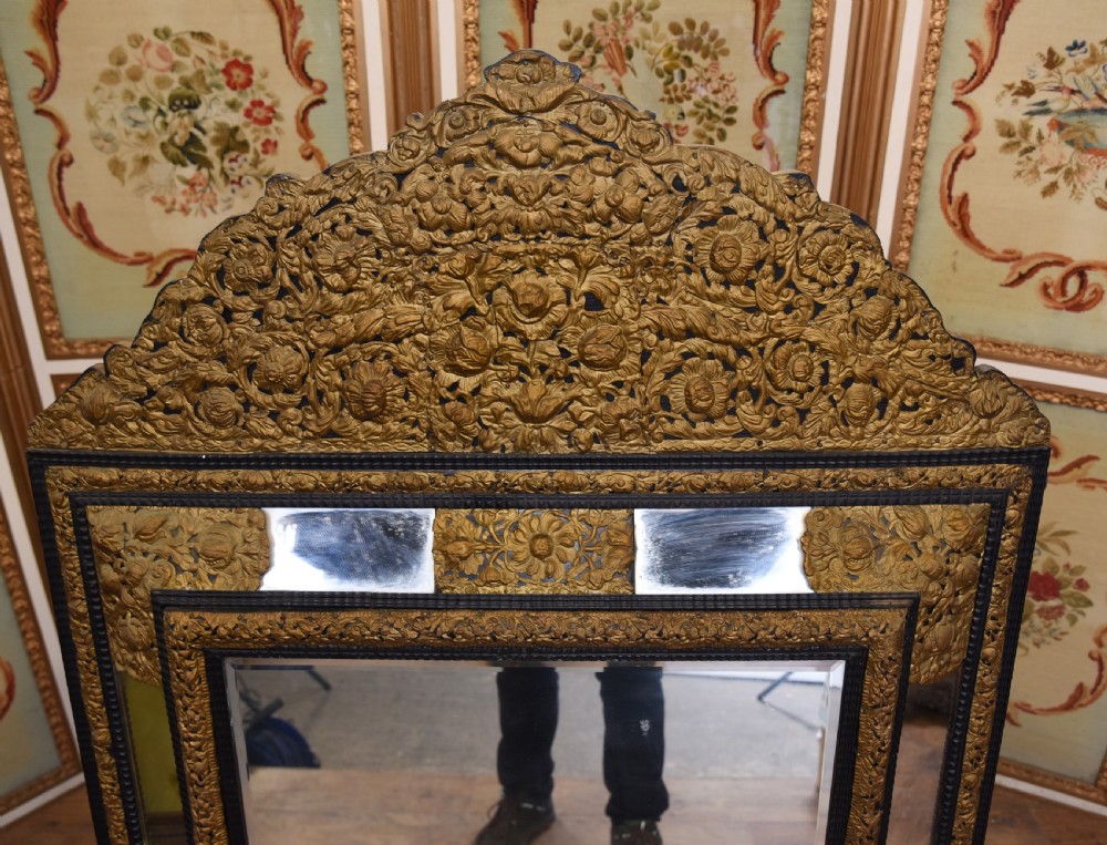 An antique French cushion mirror featuring an ornate repousse brass crest, detailed floral metal mounts, and a multi-panelled glass frame. View V