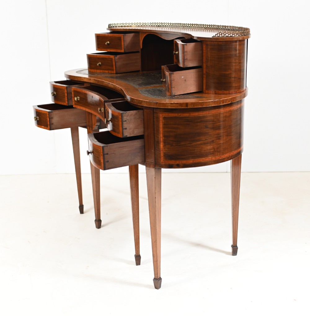 An Edwardian mahogany kidney-shaped writing desk featuring a brass gallery top, inlaid drawers, black leather writing inserts, and tapered legs. View X
