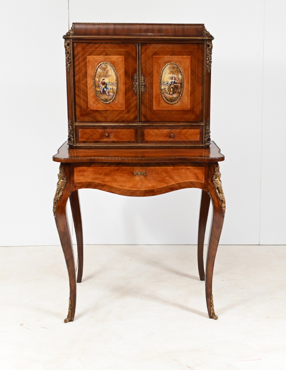 A French Louis XV style kingwood bonheur de jour desk featuring two upper cabinet doors inset with oval painted porcelain plaques, ornate metal mounts, and cabriole legs.