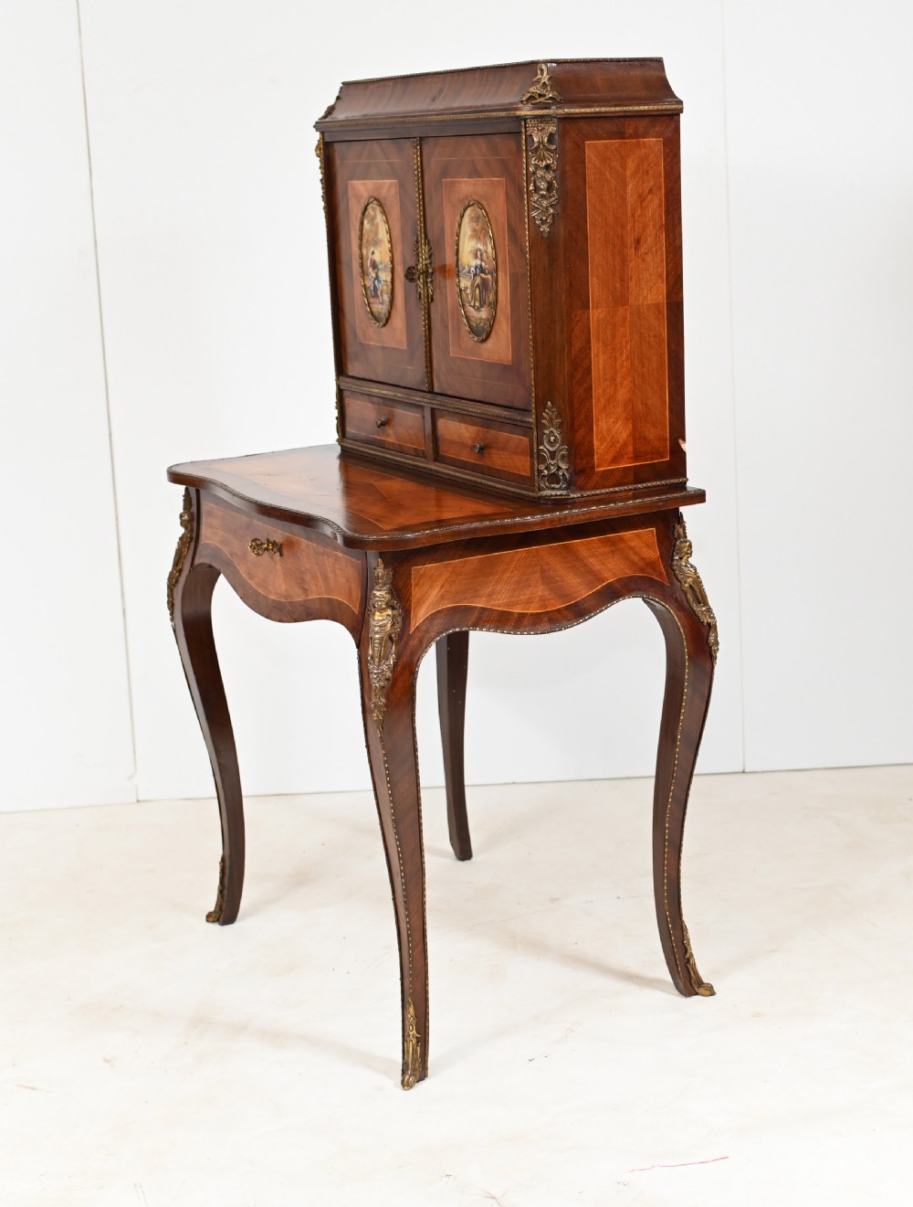 A French Louis XV style kingwood bonheur de jour desk featuring two upper cabinet doors inset with oval painted porcelain plaques, ornate metal mounts, and cabriole legs. View VII