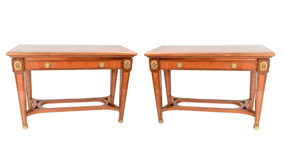 A pair of French Empire style mahogany console tables featuring satinwood crossbanding, gilt-bronze rosette mounts, and curved lower stretchers.