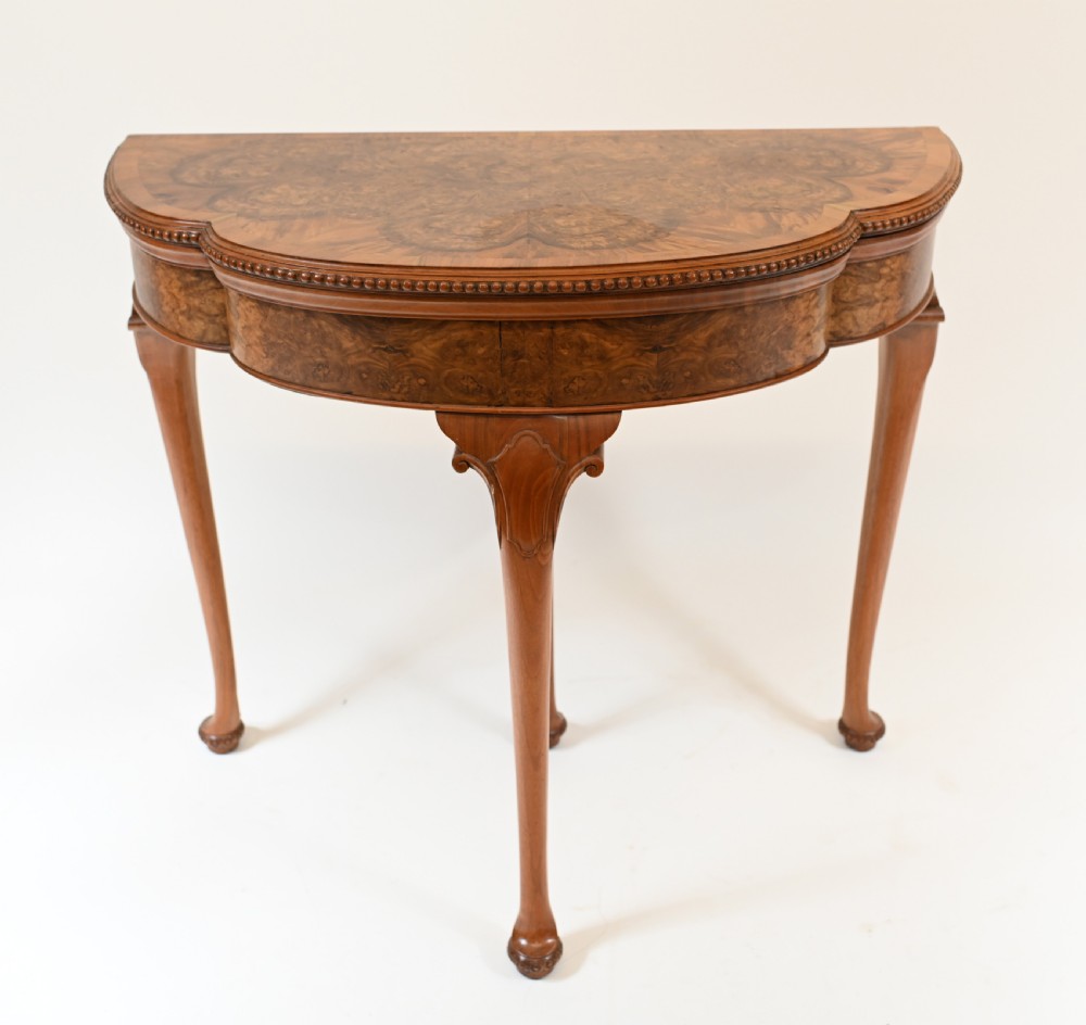 A Queen Anne style burr walnut fold-over card table featuring a scalloped demilune top, bead moulding, and four cabriole legs ending in pad feet.