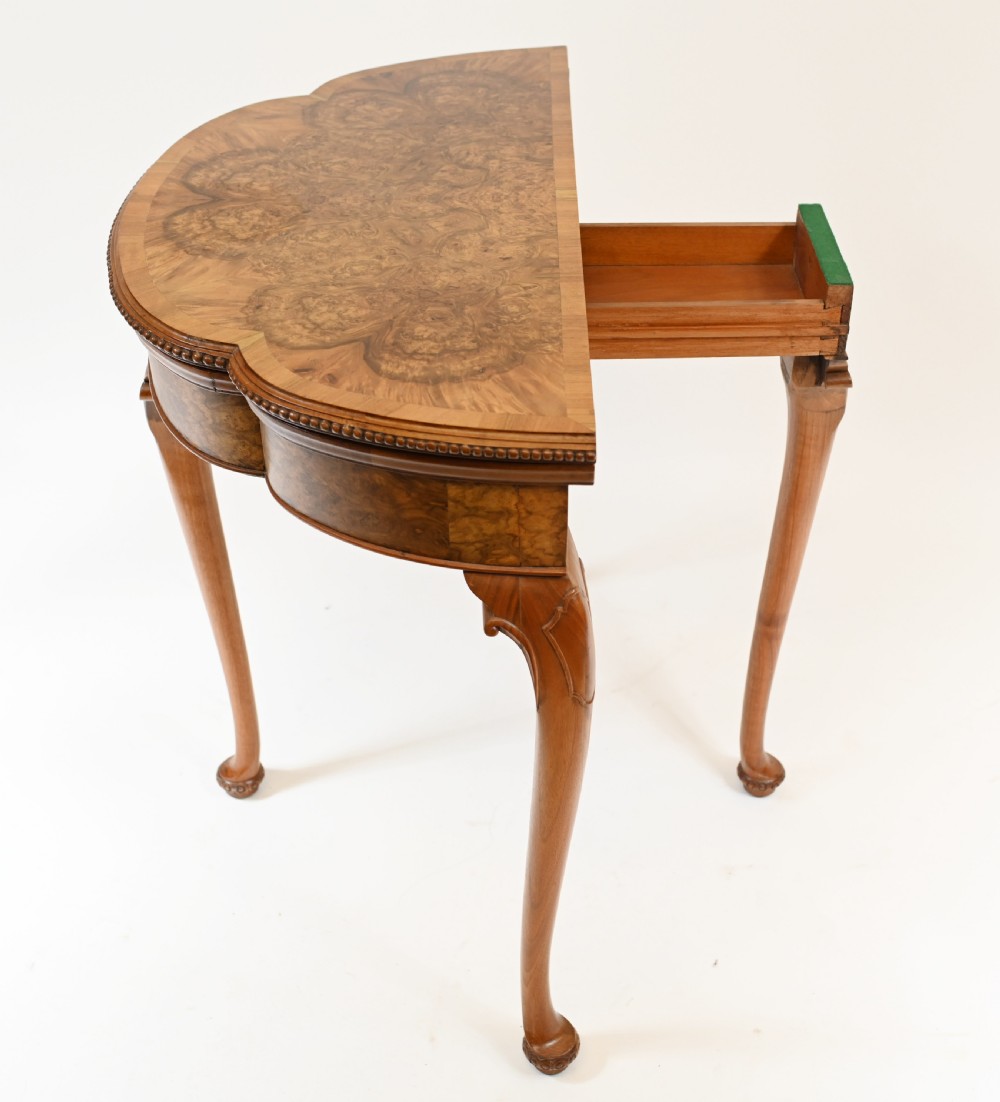 A Queen Anne style burr walnut fold-over card table featuring a scalloped demilune top, bead moulding, and four cabriole legs ending in pad feet. View V