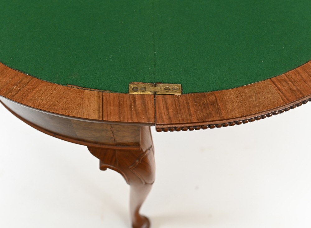 A Queen Anne style burr walnut fold-over card table featuring a scalloped demilune top, bead moulding, and four cabriole legs ending in pad feet. View VII