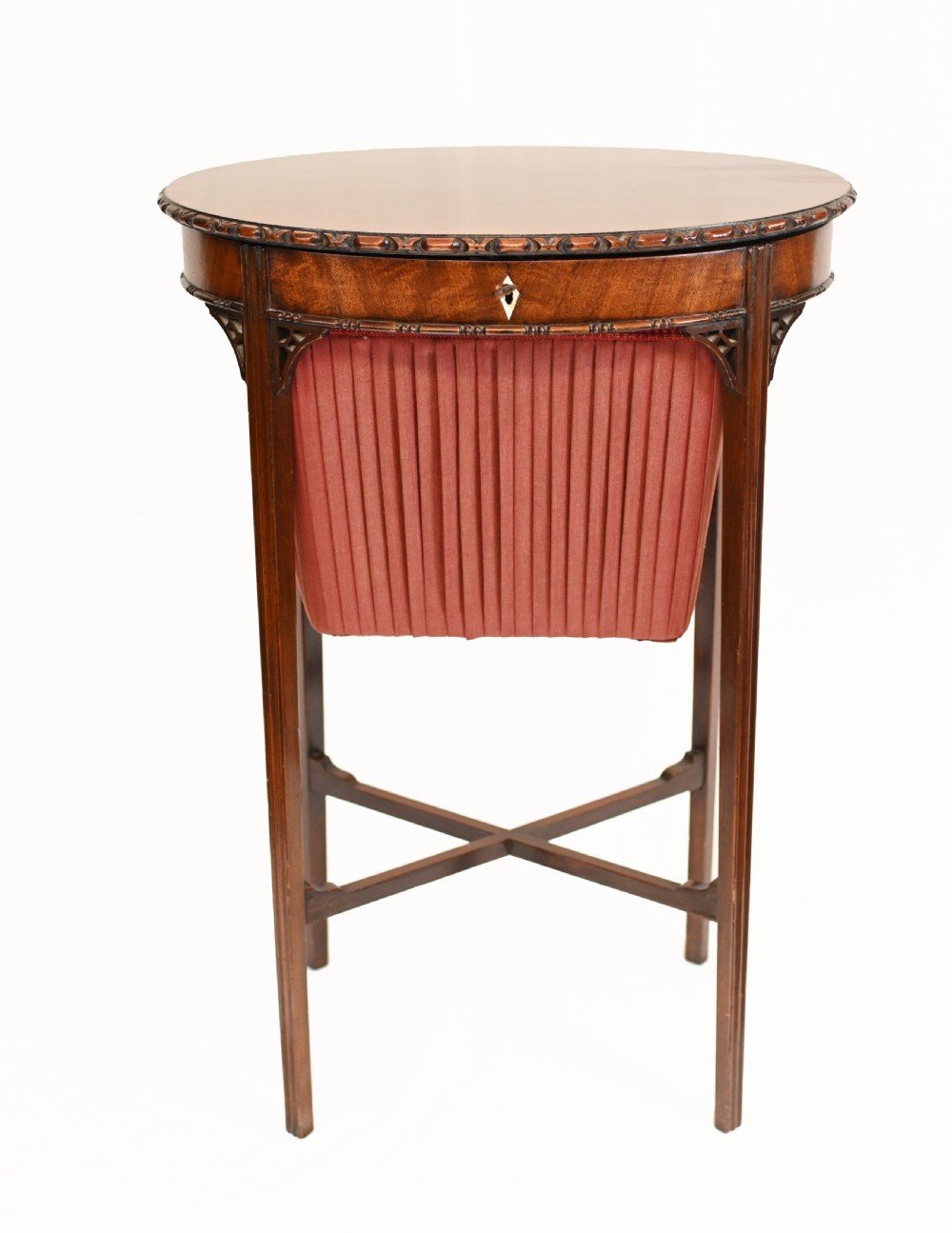 An Edwardian mahogany sewing table featuring a carved top rim, a central keyhole escutcheon, a pleated red fabric basket underneath, and tapered legs connected by an X-frame stretcher.
