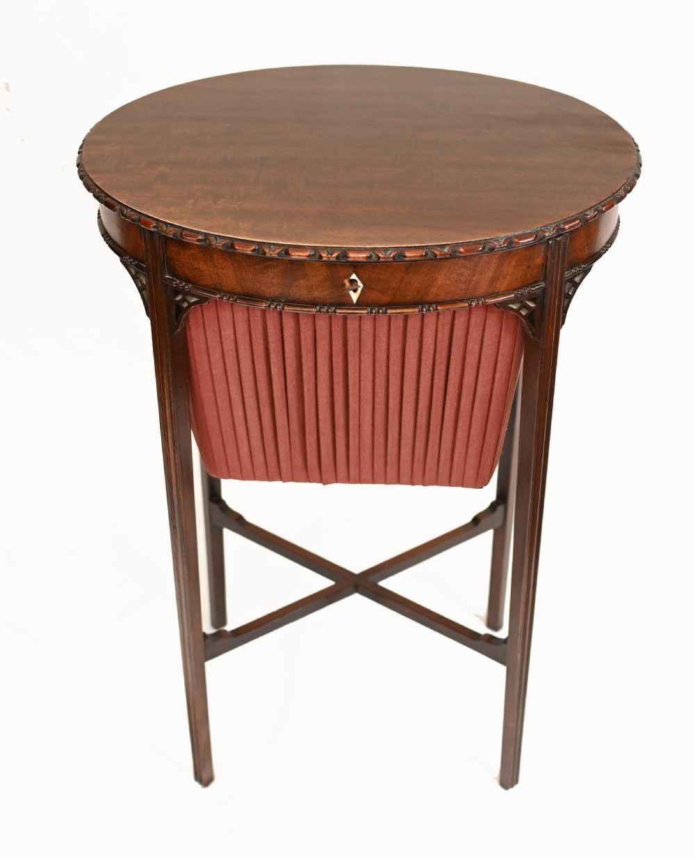 An Edwardian mahogany sewing table featuring a carved top rim, a central keyhole escutcheon, a pleated red fabric basket underneath, and tapered legs connected by an X-frame stretcher. View III
