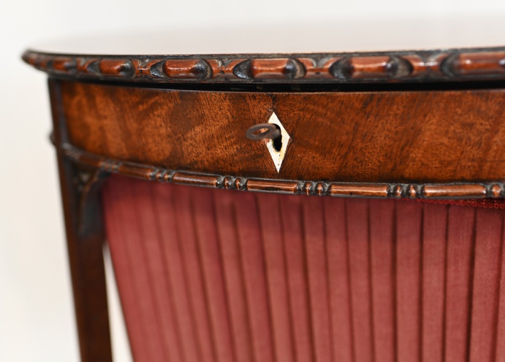 An Edwardian mahogany sewing table featuring a carved top rim, a central keyhole escutcheon, a pleated red fabric basket underneath, and tapered legs connected by an X-frame stretcher. View IV