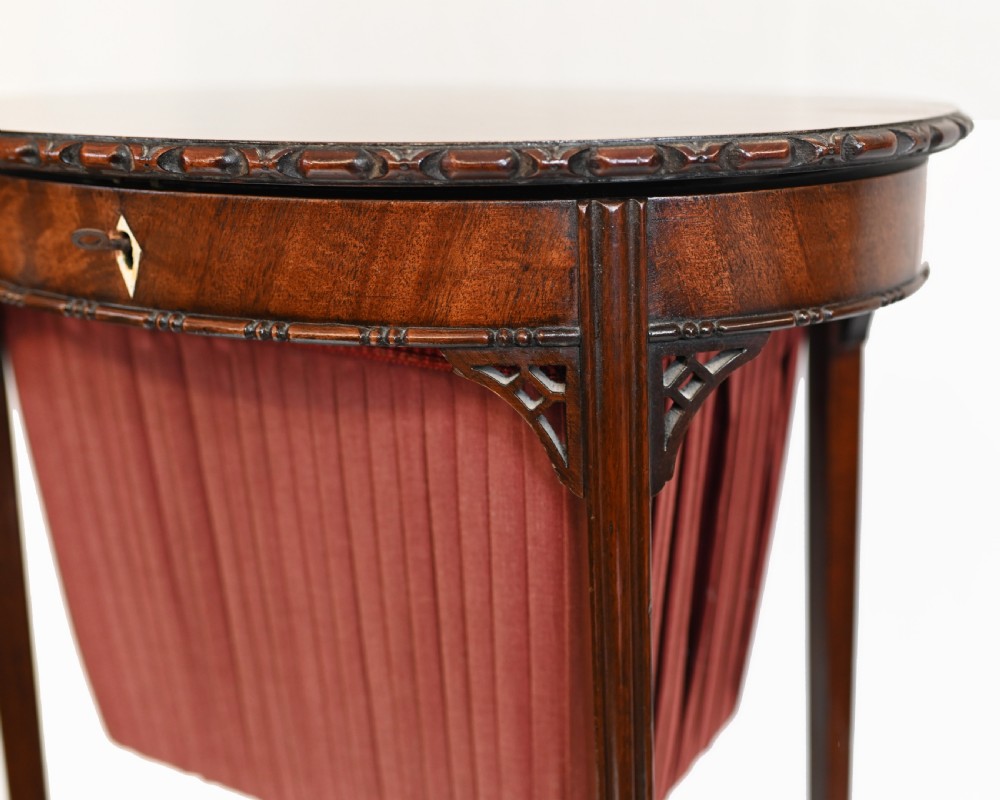 An Edwardian mahogany sewing table featuring a carved top rim, a central keyhole escutcheon, a pleated red fabric basket underneath, and tapered legs connected by an X-frame stretcher. View V