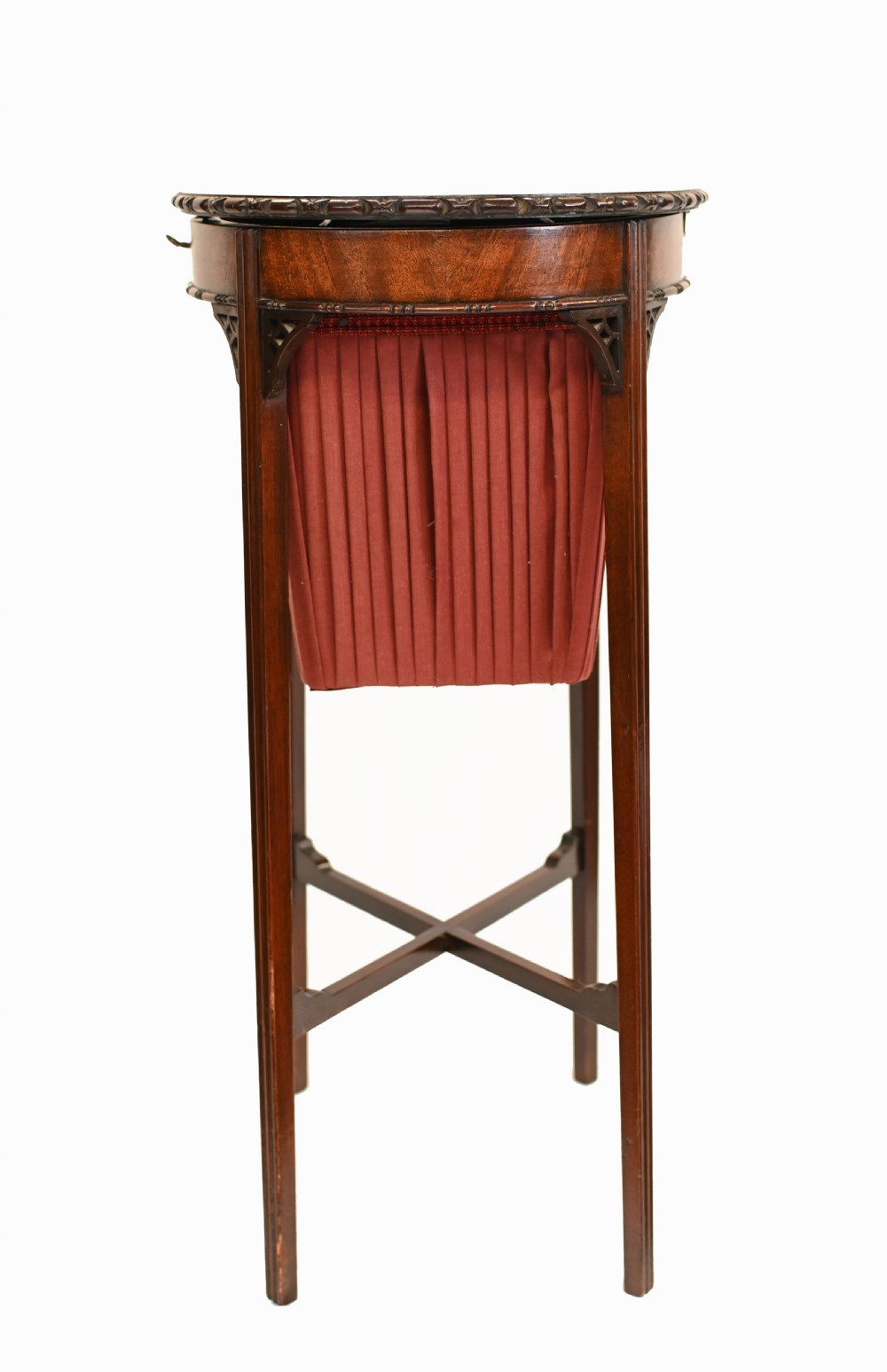 An Edwardian mahogany sewing table featuring a carved top rim, a central keyhole escutcheon, a pleated red fabric basket underneath, and tapered legs connected by an X-frame stretcher. View VI