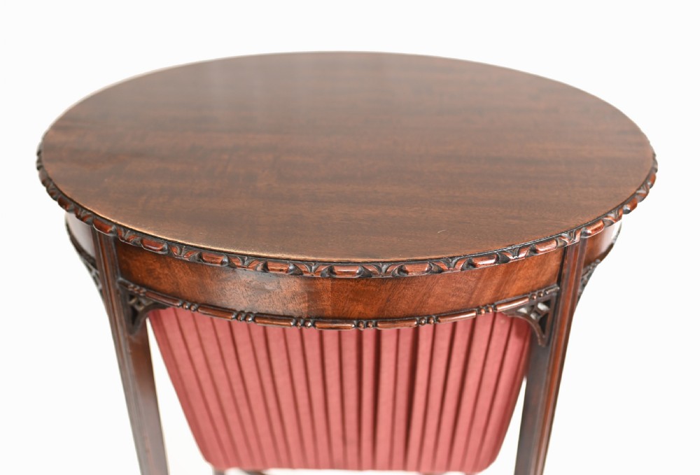 An Edwardian mahogany sewing table featuring a carved top rim, a central keyhole escutcheon, a pleated red fabric basket underneath, and tapered legs connected by an X-frame stretcher. View VII
