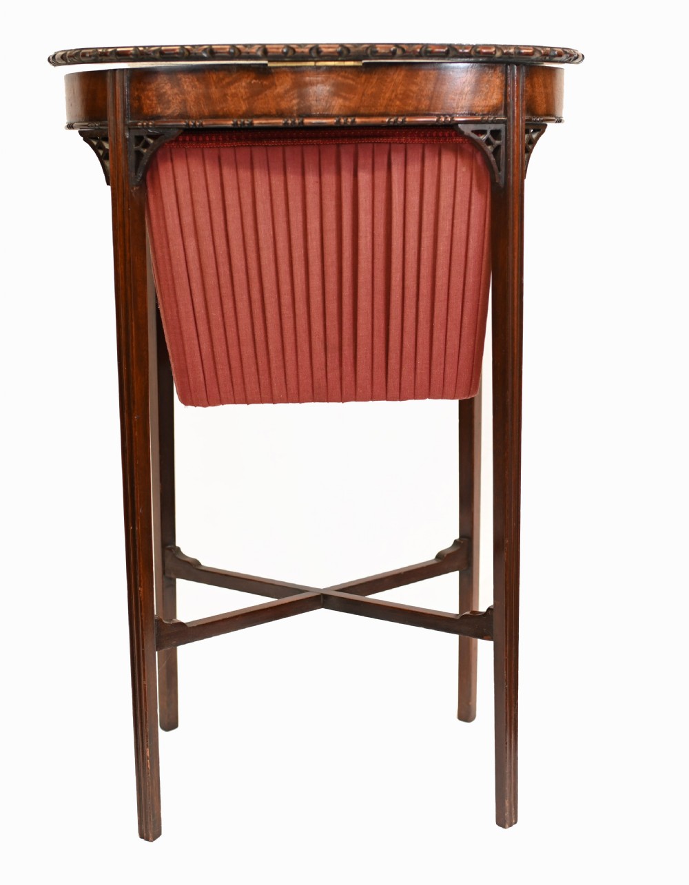 An Edwardian mahogany sewing table featuring a carved top rim, a central keyhole escutcheon, a pleated red fabric basket underneath, and tapered legs connected by an X-frame stretcher. View VIII