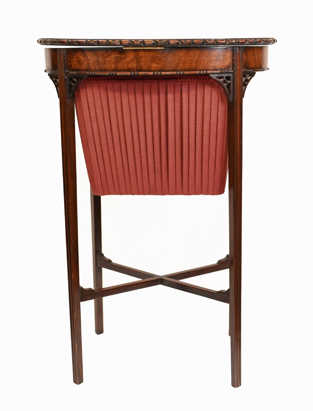 An Edwardian mahogany sewing table featuring a carved top rim, a central keyhole escutcheon, a pleated red fabric basket underneath, and tapered legs connected by an X-frame stretcher. View IX
