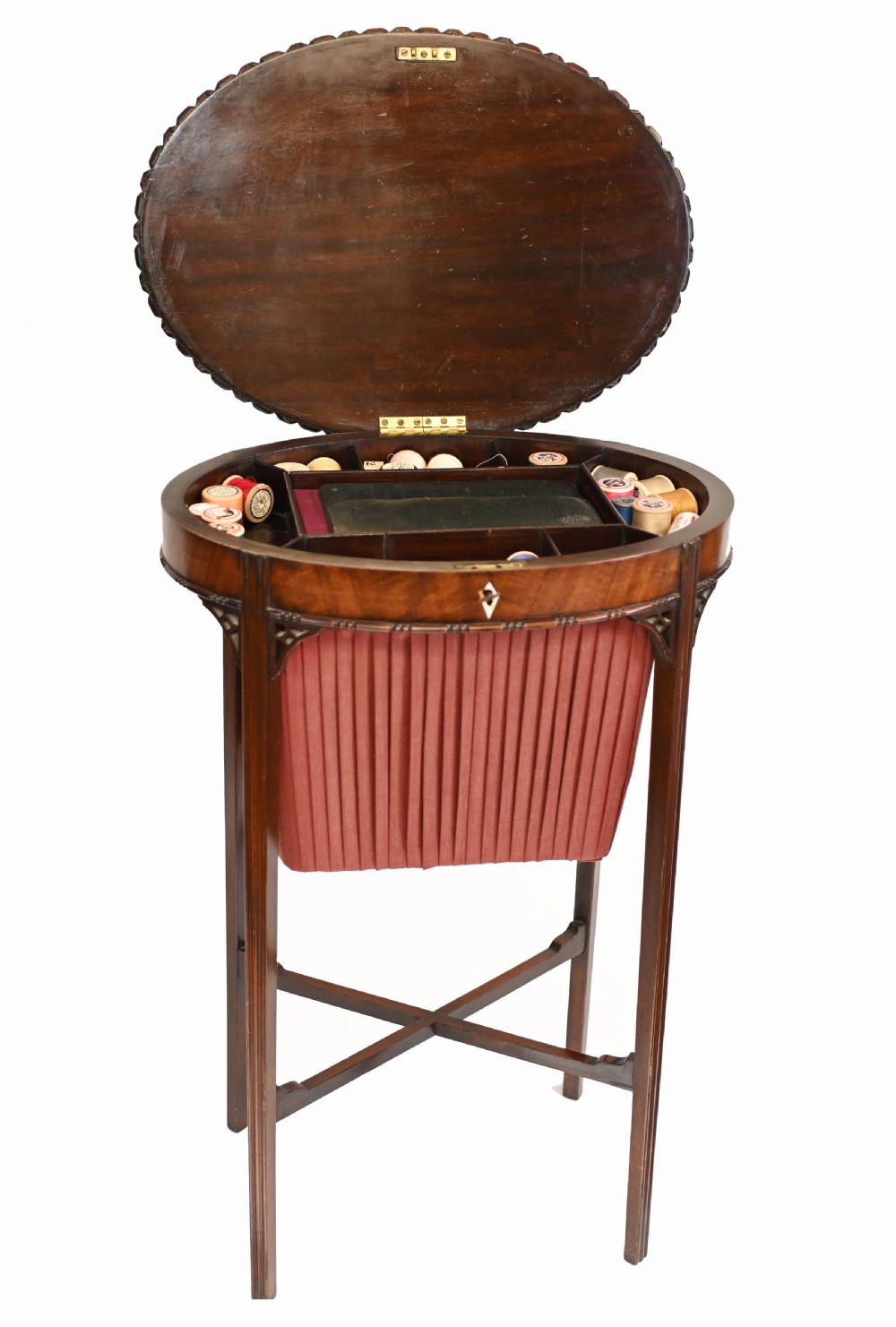 An Edwardian mahogany sewing table featuring a carved top rim, a central keyhole escutcheon, a pleated red fabric basket underneath, and tapered legs connected by an X-frame stretcher. View X