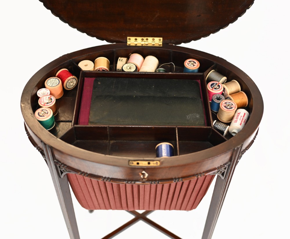 An Edwardian mahogany sewing table featuring a carved top rim, a central keyhole escutcheon, a pleated red fabric basket underneath, and tapered legs connected by an X-frame stretcher. View XI