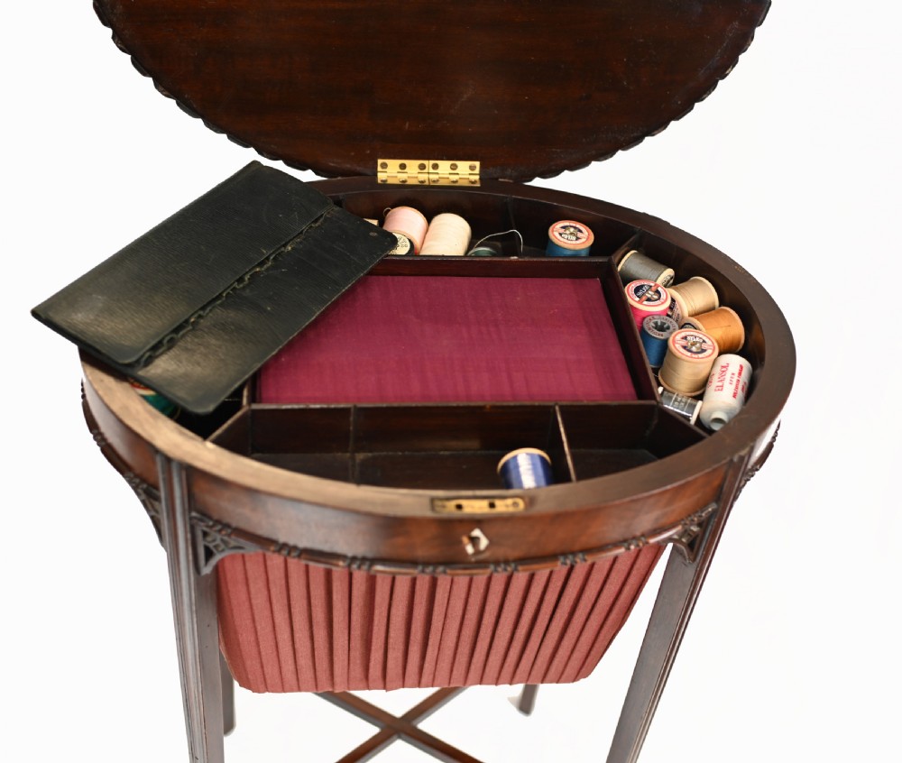 An Edwardian mahogany sewing table featuring a carved top rim, a central keyhole escutcheon, a pleated red fabric basket underneath, and tapered legs connected by an X-frame stretcher. View XII