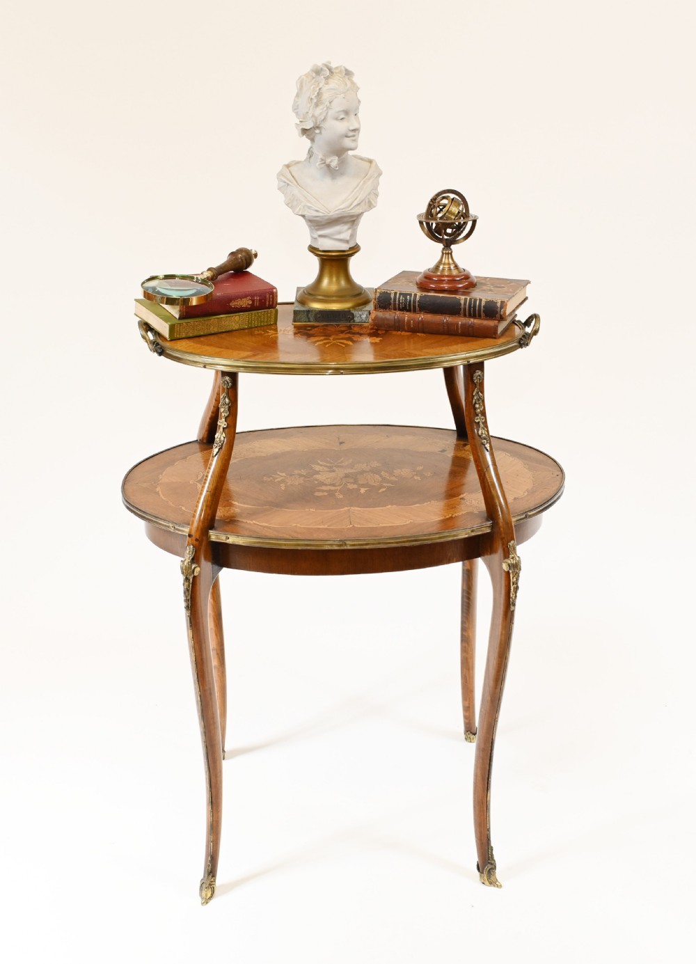 An antique French two-tiered oval etagere side table featuring floral marquetry inlay, slender cabriole legs, and polished brass mounts.