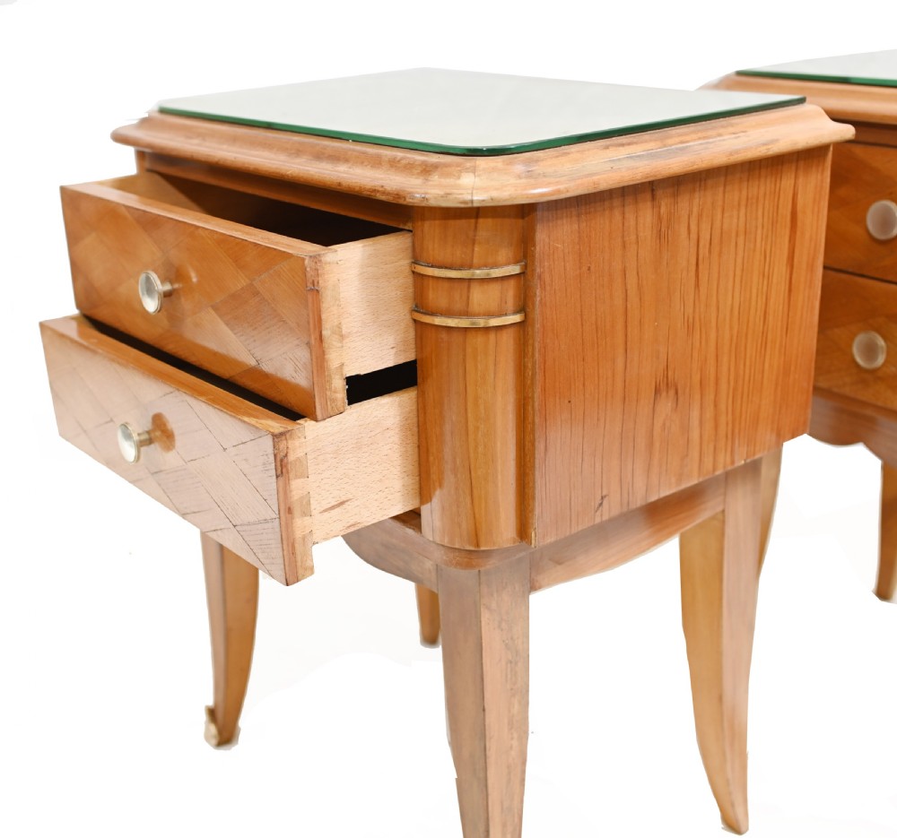 A pair of 1930s Art Deco wooden bedside cabinets featuring two drawers with diamond geometric veneers, rounded top edges with glass surface protectors, brass accents, and tapered legs. View V