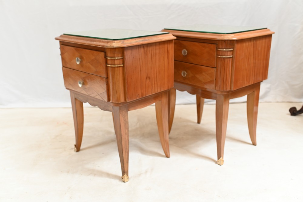 A pair of 1930s Art Deco wooden bedside cabinets featuring two drawers with diamond geometric veneers, rounded top edges with glass surface protectors, brass accents, and tapered legs. View VI