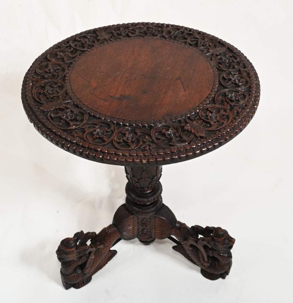 A dark brown carved timber side table featuring a circular top with a pierced floral border, supported on a turned stem and tripod legs shaped like mythological beasts.