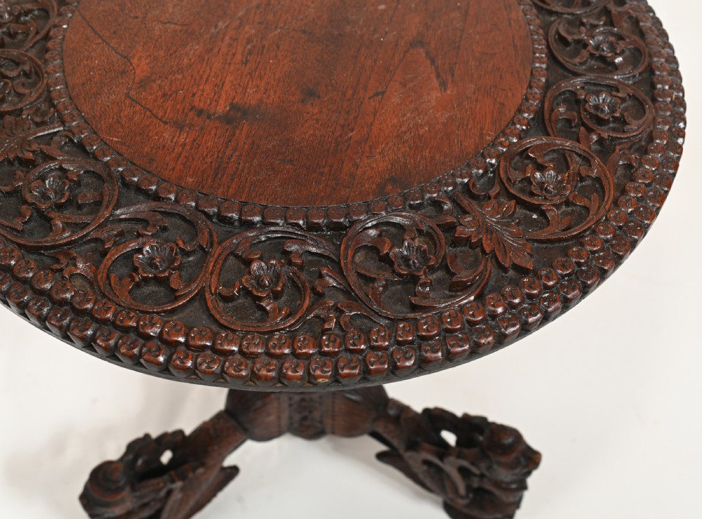 A dark brown carved timber side table featuring a circular top with a pierced floral border, supported on a turned stem and tripod legs shaped like mythological beasts. View II