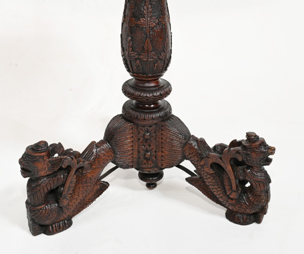 A dark brown carved timber side table featuring a circular top with a pierced floral border, supported on a turned stem and tripod legs shaped like mythological beasts. View IV