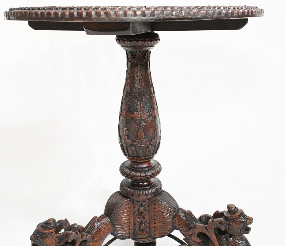A dark brown carved timber side table featuring a circular top with a pierced floral border, supported on a turned stem and tripod legs shaped like mythological beasts. View V