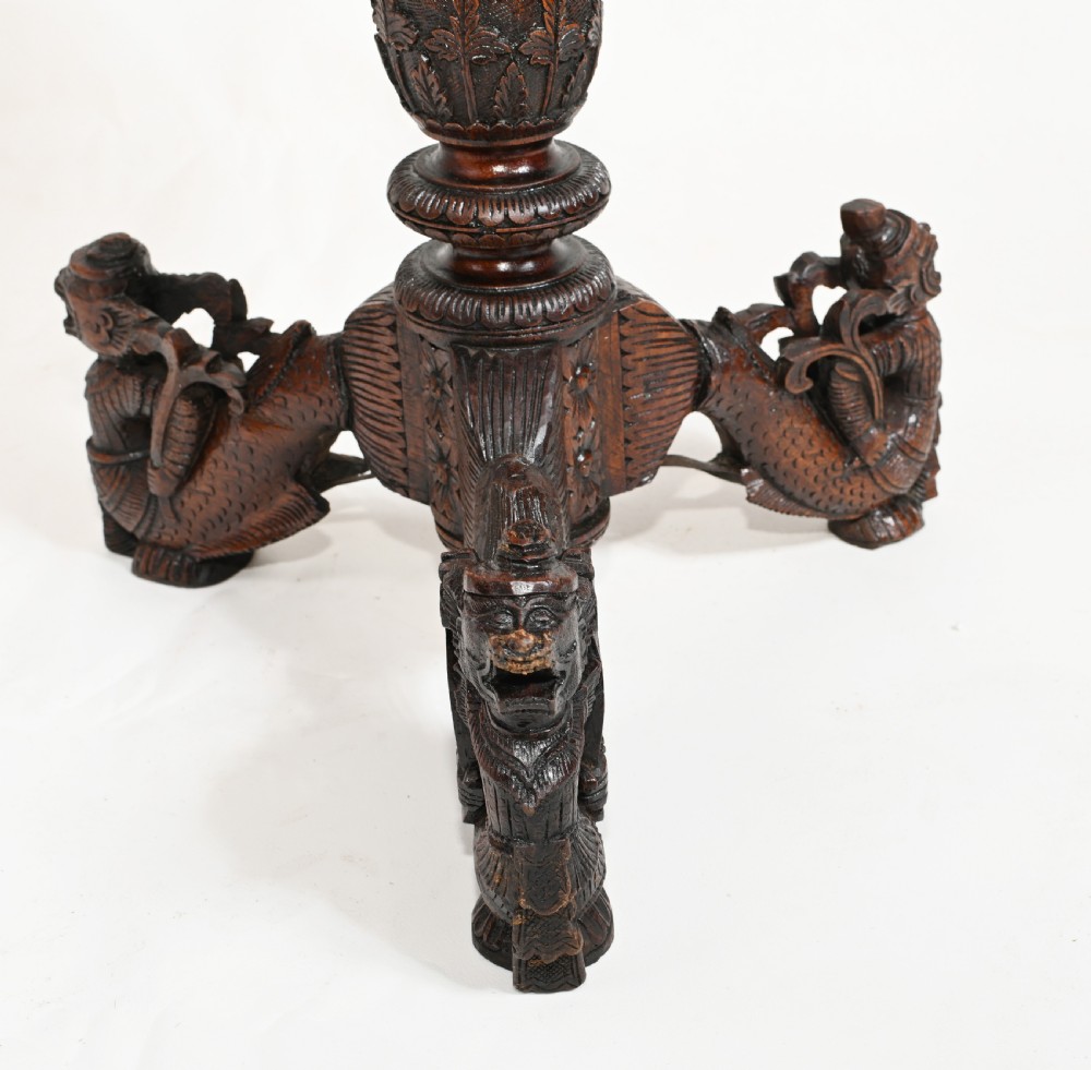 A dark brown carved timber side table featuring a circular top with a pierced floral border, supported on a turned stem and tripod legs shaped like mythological beasts. View VI